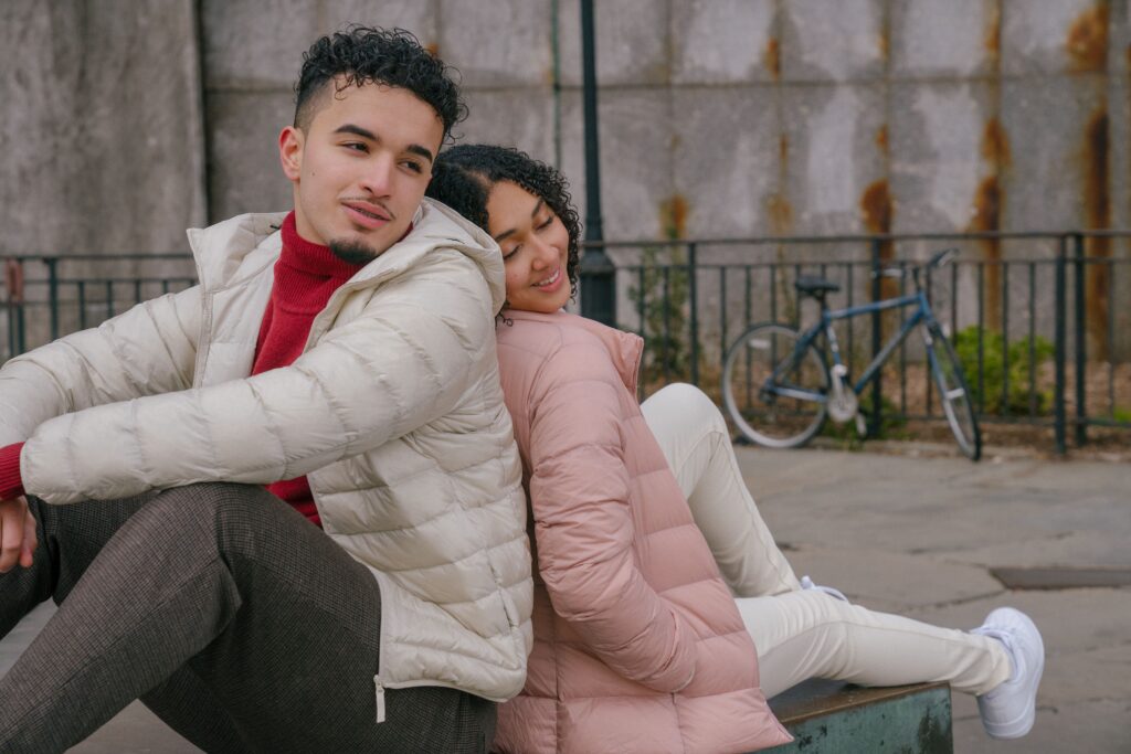 Happy young ethnic couple in stylish clothes sitting on bench back to back while resting on street